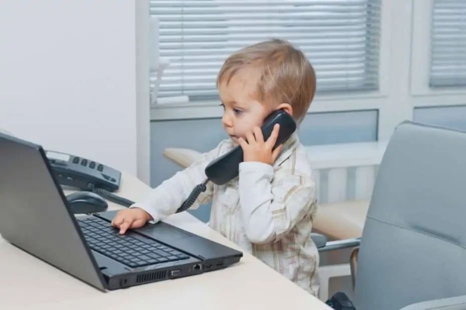 Mitbringen eines Kindes ins Büro: Das mitgebrachte Kleinkind sitzt auf dem Schreibtischstuhl im Büro, hält den Telefonhörer und tippt auf dem Laptop.